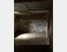 Ein dunkler, leerer Kellerraum mit teils abblätterndem Putz an den Wänden, einer Ziegelmauer auf der rechten Seite und einem Boden aus Ziegelsteinen. Licht fällt schwach von links in den Raum und hebt die raue Struktur der Wände hervor.