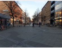 Ein städtischer Platz mit Kopfsteinpflaster und mehreren Reihen von Fahrrädern und E-Scootern an den Seiten. Im Hintergrund sind mehrstöckige Wohn- und Geschäftsgebäude, darunter ein Apothekenladen, sowie kahler Bäume zu sehen.