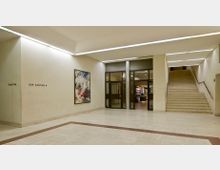 Ein heller Innenbereich mit einem Marmorboden, Treppen auf der rechten Seite und großen Glastüren in der Mitte. An der Wand hängt ein modernes Kunstwerk, und links steht „Zur Kantine“ als Wegweiser.