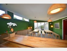 Gemütlicher Innenraum mit grünen Wänden, Holztischen und Bänken in minimalistischem Design. Große Fenster und Glastür lassen Tageslicht herein, ergänzt durch hängende Rattanlampen und Pflanzen als Dekoration.