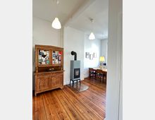 Wohnzimmer mit Holzdielenboden, links ein rustikales Holzbuffet mit Dekorationen, in der Mitte ein freistehender Kaminofen auf einer Glasplatte, im Hintergrund ein Esstisch mit Stühlen und Pendelleuchten.