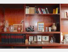 Holzregal mit mehreren Einlegeböden, die verschiedene dekorative Objekte wie Bücher, Vasen, Glasgefäße, einen kleinen Kerzenhalter, Bilderrahmen und Skulpturen präsentieren. Im unteren Teil sind gebundene Bücher mit roten Titeln auf schwarzem Einband zu sehen.