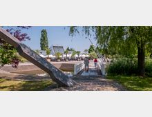 Ein Außenbereich mit einer kleinen Brücke, die über ein Wasserbecken führt, umgeben von üppiger Vegetation und Bäumen. Im Hintergrund sind ein modernes Gebäude mit Glasdach, Palmen in Kübeln und Sitzbereiche mit Sonnenschirmen zu sehen.