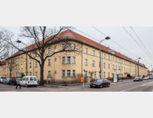 Gelbes Wohngebäude mit roten Ziegeldächern in einer Straßenecke, links von einem großen Baum verdeckt. Vor dem Gebäude befinden sich eine Bushaltestelle, geparkte Autos und eine Straßenbahntrasse mit Oberleitungen.