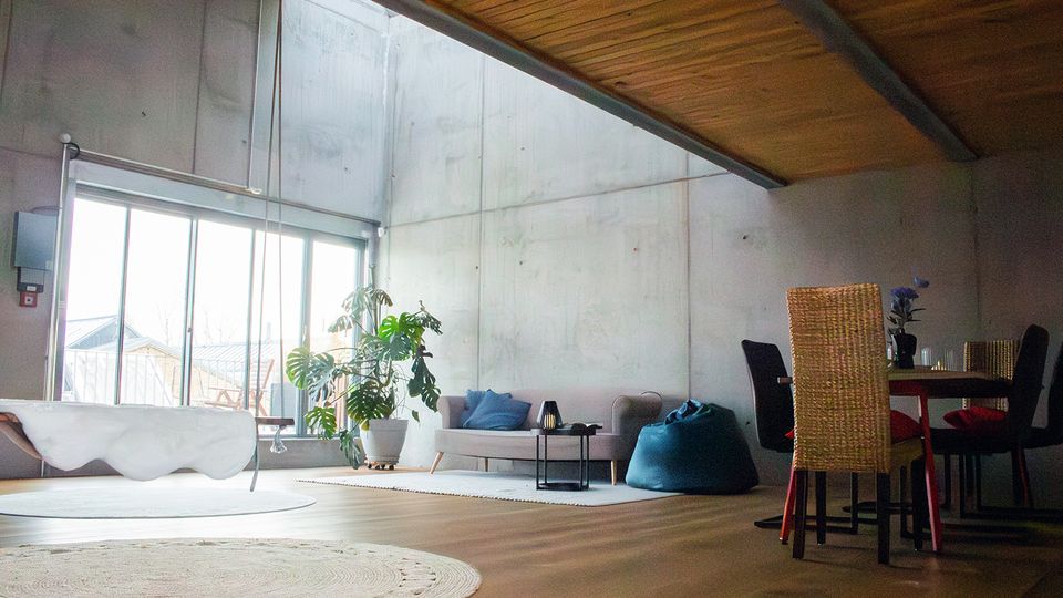 Helles Loft-Wohnzimmer mit hohen Betonwänden und schräger Holzdecke, großen Schiebetüren zur Terrasse und viel Tageslicht. Im Raum stehen eine freistehende Badewanne, Sofa mit Beistelltisch, Sitzsack, große Zimmerpflanze sowie ein Essbereich mit Tisch und Stühlen.