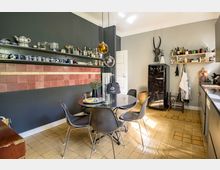 Eine stilvolle Küche mit dunklen Wänden und einem roten Fliesenspiegel. Ein runder Tisch mit schwarzen Stühlen steht in der Mitte, darüber hängen moderne Pendelleuchten, während offene Regale mit Geschirr und Vorräten dekoriert sind. Im Hintergrund steht ein schwarzer, vintage-inspirierter Kühlschrank.