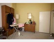 Wohnzimmer mit gelber Wand und grauem Teppichboden. Zu sehen sind ein Holzschrank, ein Bügelbrett mit Dampfbügeleisen, eine Stehlampe, eine Kommode mit verschiedenen Gegenständen und eine angelehnte Zimmertür.