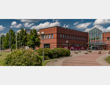 Backstein-Gebäude eines Einkaufszentrums mit der Aufschrift "Oder-Center", einem gläsernen Eingangsbereich und umliegenden Grünflächen sowie Laternen. Im Vordergrund führt ein gepflasterter Weg auf den Eingang zu, flankiert von kleinen Büschen und Bäumen.