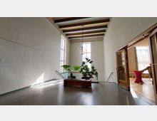 Offener, heller Innenraum mit hohen Decken, großen Fenstern und sichtbaren Holzdeckenbalken. In der Mitte steht eine Bank vor einer Glasbalustrade mit Topfpflanzen, daneben führen zwei Treppen hinunter. Rechts ist eine offene Tür zu einem Raum mit Parkettboden und Stehtischen.