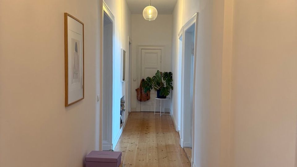 Long, narrow residential hallway with white walls, a light wood plank floor, and a shallow arched ceiling near the foreground. A globe pendant light hangs overhead, with doorways along both sides, a wooden slatted bench and framed wall art on the left, and potted plants near a closed door at the far end.
