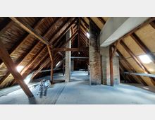 Dachboden eines Gebäudes mit sichtbarer Holzbalkenkonstruktion, mehreren Stützpfeilern aus Holz und Mauerwerk sowie einem Boden aus Holzplanken. Seitliche Dachfenster lassen Tageslicht in den Raum.