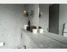 Moderne Badezimmergestaltung mit einer grauen Steinoberfläche und größtenteils spiegelnder Fläche. Auf einer Ablage stehen ein dunkles Glas mit Blumen, gefaltete Handtücher und kleine Boxen; im Hintergrund sind ein Handtuchhalter und weitere graue Steinwände sichtbar.