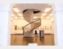 Innenansicht eines Ausstellungsraums mit weißen Wänden, beleuchtet von Deckenlampen. Im Mittelpunkt steht eine Wendeltreppe aus Beton mit einem Metallgeländer, umgeben von Kunstwerken, die an den Wänden hängen.