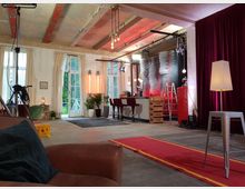 Offener Raum im Loft-Stil mit rostikaler Decke und großen Fenstern, die mit hellen Vorhängen dekoriert sind. Im Raum befinden sich eine Couch, eine Bar mit hohen Stühlen, Pflanzen, Studiobeleuchtung sowie ein roter Teppich, Kameraequipment und eine rote Kühlschrankkulisse.