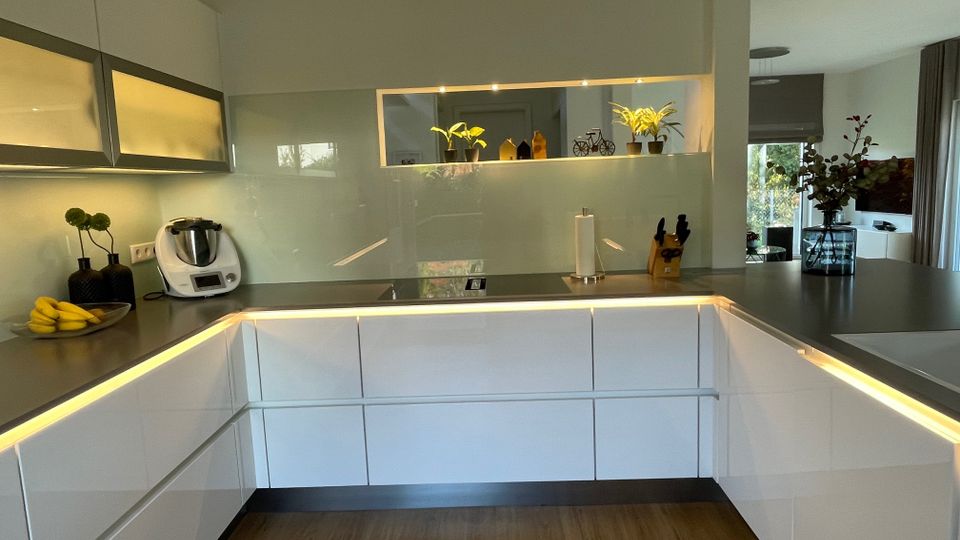 Eine moderne Küche mit weißen, schlichten Fronten und einer dunklen Arbeitsplatte. Eine indirekte Beleuchtung unter den Schränken und Regalen sorgt für Akzente; der Raum enthält Deko-Elemente wie Pflanzen, ein Messerblock, und eine Küchenmaschine auf der Arbeitsfläche.
