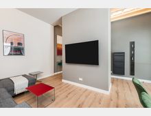 Modernes Wohnzimmer mit hellen Wänden und Holzfußboden, einer grauen Sofaecke, Glastisch mit roter Ablage und einem großen wandmontierten Flachbildfernseher. An der Wand hängen gerahmte Kunstposter; rechts ist ein Durchgang zu einem weiteren Bereich mit dunklen Wandpaneelen und eingebauten Elementen zu sehen.