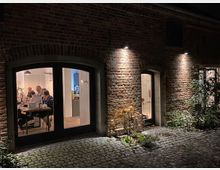 Backsteinfassade eines Gebäudes mit drei beleuchteten Rundbogenfenstern; zwei der Fenster geben Einblick in einen Innenraum, wo Menschen an einem Tisch sitzen. Der Boden vor dem Gebäude besteht aus Kopfsteinpflaster, und einige Pflanzen säumen den Eingang.