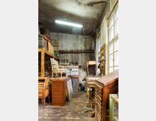Eine alte Werkstatt mit verwitterter Decke und Holzboden, gefüllt mit Vintage-Möbeln, Werkzeugen an der Wand und Schubladenschränken. Das natürliche Licht kommt durch große Fenster auf der rechten Seite, während eine Neonleuchte die Decke erhellt.
