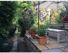 Gepflegte Gartenterrasse mit Pflastersteinen und einer niedrigen Natursteinmauer, umgeben von dichtem Grün. Auf der Terrasse stehen Blumentöpfe, ein Holztisch mit Sitzbänken und ein Sonnenschirm; im Hintergrund sind Bäume und ein kleiner Gartenweg erkennbar.