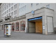 Eingang zum U-Bahnhof Frankfurter Tor in Berlin, mit einem blauen Schild und der Beschriftung "U-Bhf Frankfurter Tor". Im Hintergrund ein historisches Gebäude mit heller Fassade, roten Fensterrahmen und Arkaden.