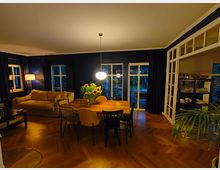 Ein Wohnzimmer mit dunklen Wänden, einem hölzernen Esstisch mit gemischten Stühlen und einem Blumenstrauß aus Sonnenblumen in der Mitte. Im Hintergrund befinden sich eine gelbe Couch, eine Stehlampe, hohe Fenster mit Vorhängen und eine Glastür, die auf eine Terrasse führt; rechts steht ein Schrank mit Geschirr.