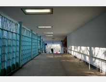 Überdachter Durchgang mit schrägem Boden, links eine blaue Metallgitterwand, rechts eine weiße Wand mit Handlauf. Im Hintergrund eine Öffnung, durch die Tageslicht einfällt, und einige Fahrräder, die an der Wand geparkt sind.