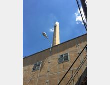 Industriegebäude mit einer beige-farbenen Fassade, kleinen Fenstern und Metallverstrebungen, im Hintergrund ein hoher Industrieschornstein unter klarem, blauem Himmel. Im Vordergrund ist eine Straßenlaterne und ein Teil einer Metalltreppe sichtbar.
