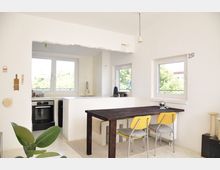 Minimalistisch gestalteter Wohnraum mit integrierter Küche, weißen Wänden und großen Fenstern, die viel Tageslicht hereinlassen. Im Vordergrund steht ein dunkler Holztisch mit gelben Stühlen, im Hintergrund sind ein Herd, eine Spüle und eine Arbeitsfläche zu sehen.