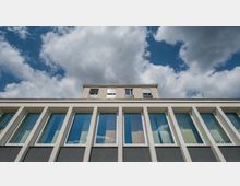 Flaches modernes Gebäude mit großen, symmetrisch angeordneten Glasfenstern im unteren Teil und einer oberen Etage mit hellen Fassadenplatten und kleinen Fenstern. Der Himmel darüber ist bewölkt mit einzelnen blauen Stellen.