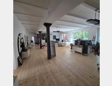 Großer, offener Raum mit Holzboden, weißen Wänden und sichtbaren weißen Deckenbalken. Mehrere schwarze Metallstützen teilen den Raum, der mit Sitzmöbeln, Schreibtischen, Regalen und großen Fenstern mit Blick auf grüne Bäume ausgestattet ist.