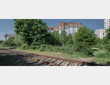 Bahnstrecke mit Schotterbett im Vordergrund, dahinter Grünflächen mit Bäumen und Büschen sowie mehrstöckige Wohngebäude mit bunter Fassade im Hintergrund. Der Himmel ist klar und blau.