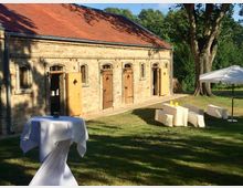 Ein langgezogenes, historisches Gebäude aus gelbem Backstein mit roten Ziegeldach und mehreren Holztüren, umgeben von einer grünen Wiese. Im Vordergrund stehen ein Stehtisch mit weißer Tischdecke, mehrere mit weißen Tüchern bedeckte Bänke sowie ein Sonnenschirm unter Bäumen.