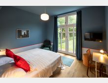 Schlafzimmer mit blauen Wänden und Holzboden, ausgestattet mit einem Doppelbett mit weißer Bettwäsche und roten Kissen, einem Schreibtisch mit Stuhl und Lampe, einem Flachbildfernseher an der Wand, einem grünen Sessel und einer großen Terrassentür, die in einen Garten führt.