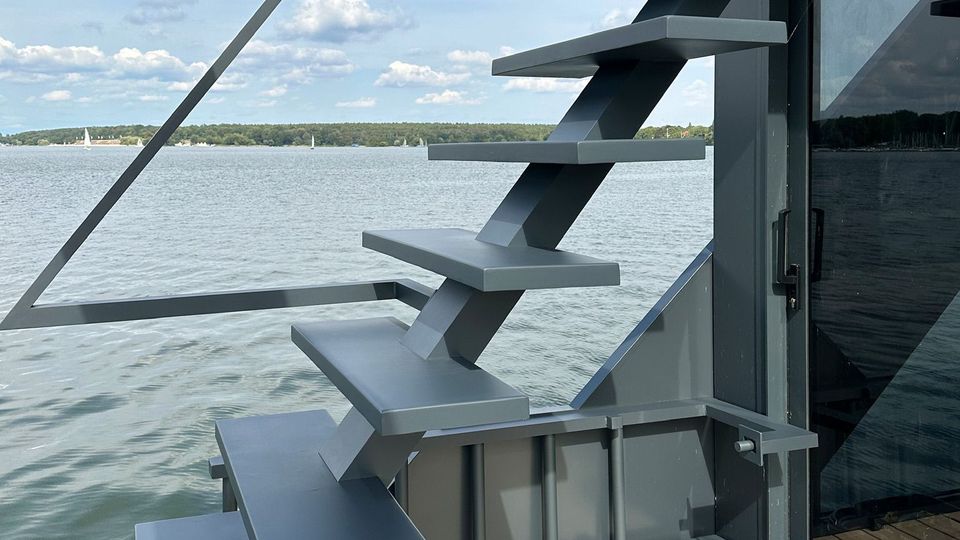 Moderne, freistehende Metalltreppe mit geradlinigem Design auf einer Terrasse aus Holzplanken, mit Blick auf einen See mit ruhiger Wasseroberfläche und bewölktem Himmel im Hintergrund.