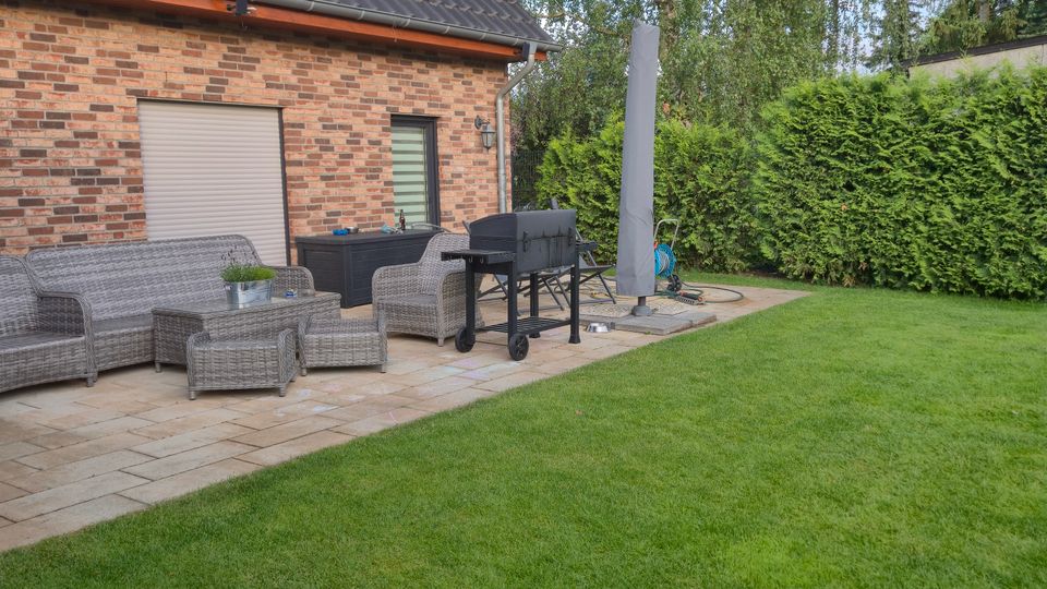 Terrasse eines Einfamilienhauses mit roter Klinkerfassade und dunkel gedecktem Satteldach mit Solarpanel. Vor der Hauswand stehen Rattan-Loungemöbel und ein Grill auf Steinplatten; daneben liegt ein gepflegter Rasen mit hoher Hecke und großen Bäumen im Hintergrund.