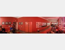 Langer, roter Innenraum eines Gebäudes mit Sitzgelegenheiten und Wanddekorationen. An den Wänden hängen Bilder, Poster und Aushänge, während Menschen auf Stühlen sitzen und sich unterhalten; rechts sind große Fenster.