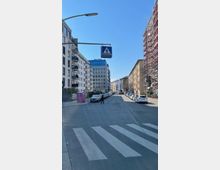 Straßenszene mit Zebrastreifen, flankiert von modernen Wohngebäuden und einem Gerüst an einem der Häuser; ein Fußgänger überquert die Straße, die von geparkten Autos gesäumt ist. Ein Verkehrszeichen für den Fußgängerüberweg hängt über der Fahrbahn, der Himmel ist klar und blau.