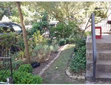 Garten mit einem schmalen, mit Steinen gesäumten Pfad, umgeben von Büschen, Blumen und Bäumen. Rechts im Bild befindet sich eine Steintreppe mit einem Metallgeländer, im Hintergrund ein geparktes Auto und ein weißer Lattenzaun.