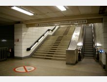 Unterirdischer U-Bahnhofeingang mit zwei Treppen: links eine klassische Steintreppe mit Handläufen und rechts daneben eine Rolltreppe. Die Wände sind mit weißen Fliesen verkleidet, und oberhalb der Treppen befinden sich Schilder mit Wegweisungen zu verschiedenen Stationen.