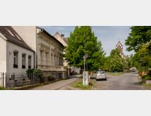 Straßenszene in einer ruhigen Wohngegend mit Kopfsteinpflaster, links eine Reihe von Häusern, darunter ein verziertes historisches Gebäude. Im Hintergrund ist eine Kirche mit markantem, neugotischen Turm zu erkennen, umgeben von grünen Bäumen.