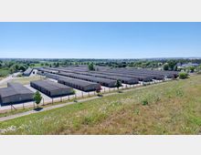 Großer Komplex aus rechteckigen, flachen Lagergebäuden mit grauen Dächern, der von einem Zaun umgeben ist. Im Hintergrund sind Wiesen, Bäume und vereinzelte Gebäude einer ländlichen Umgebung sichtbar.