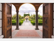 Blick durch eine große Holztür mit Rundbögen und Steinsäulen auf einen gepflegten Garten mit symmetrischen Buchsbaumhecken, einem zentralen Brunnen und einem mit Efeu bewachsenen gelben Pavillon im Hintergrund.