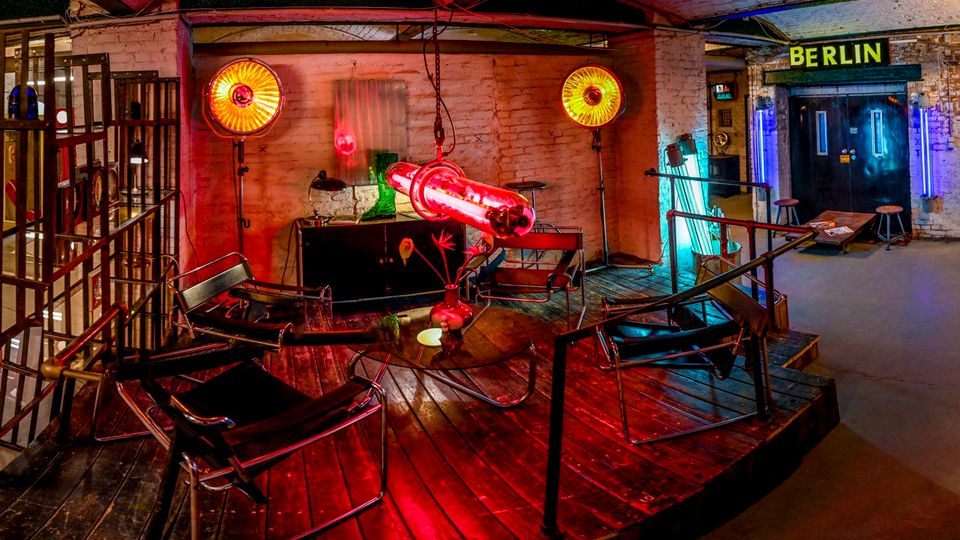 Industrieartig gestalteter Innenraum mit gemauerten Tonnengewölben, Holzpodest und schwarzen Metallstühlen um eine hängende Neonröhre; warme Scheinwerfer und farbige Lichtakzente beleuchten die Backsteinwände. Rechts ist ein Eingang mit dem Schriftzug „BERLIN“ sowie Geländern und Barhockern zu sehen.