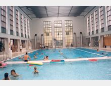 Innenansicht eines Hallenbads mit einem großen Schwimmbecken, in dem mehrere Personen schwimmen und spielen. Das Gebäude hat hohe Decken, große Fenster und eine Reihe von symmetrischen Lichtsäulen an den Wänden.