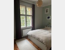 Helles Schlafzimmer mit großem Sprossenfenster und dunklen Vorhängen, darunter ein Heizkörper; draußen sind Bäume zu sehen. Ein weißes Bett mit Bettdecke steht neben einem kleinen Nachttisch, an der Wand hängen gerahmte Bilder und eine geflochtene Deckenlampe.