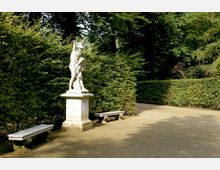 Eine Parkszene mit einer weißen Statue auf einem hohen Sockel, flankiert von zwei Steinbänken. Die Umgebung ist von gepflegten Hecken und Bäumen geprägt, die einen schattigen Weg einrahmen.