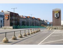 Straßenkreuzung mit zwei großen Ziegelpfeilern auf beiden Seiten, die Wappen tragen, darunter Berlins Wappen mit einem Bären. Im Hintergrund befinden sich Altbauten mit roten Dächern und ein grüner Zaun entlang der Straße.