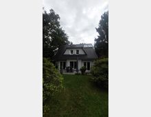 Ein kleines Einfamilienhaus mit Satteldach, umgeben von dichtem Grün und Büschen. Im Vordergrund eine gepflegte Rasenfläche, die zu einer Terrasse mit Sitzmöbeln und Blumenkübeln führt.