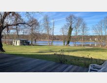 Weitläufige Rasenfläche mit vereinzelten Bäumen, im Hintergrund ein kleiner See und mehrere Häuser am Ufer; im Vordergrund eine Holzterrasse und ein Gartenpavillon auf der linken Seite. Klarer Himmel und unbelaubte Bäume deuten auf eine Winter- oder Frühjahrsaufnahme hin.