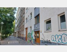 Straßenansicht mit mehrstöckigen Wohnhäusern, schlichten Fassaden und teilweise Graffiti an den Wänden. Links führt ein gepflasterter Gehweg mit Pollern und Straßenlaternen an Bäumen entlang.